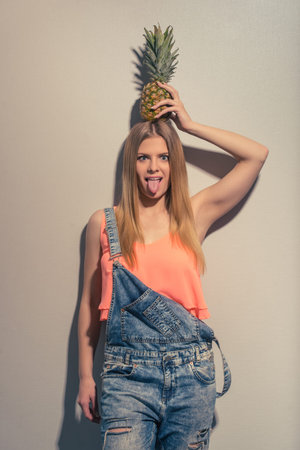 Attractive girl in summer clothes is holding a pineapple overhead, looking at camera and showing...