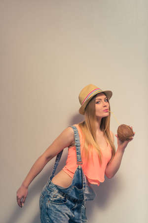 Attractive girl in summer clothes and hat is drinking a coconut milk, looking at camera and smiling, against gray backgroundの写真素材