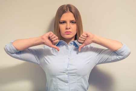 Attractive girl in classic clothes is showing not Ok sign and looking at camera, against gray...