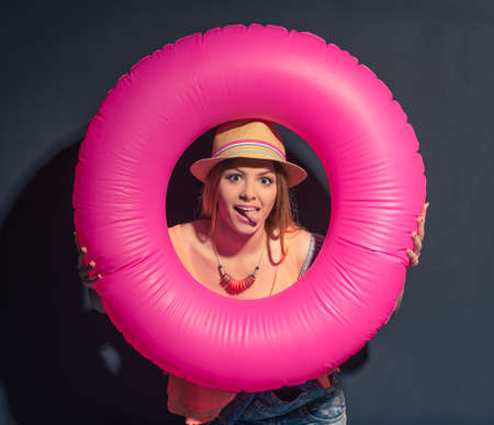 Attractive girl in summer clothes is holding a swim ring, looking through it at camera and showing tongue, against dark backgroundの写真素材