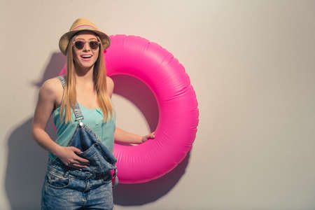 Attractive girl in summer clothes and sun glasses is holding a swim ring, looking at camera and smiling, against gray backgroundの写真素材