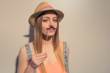 Attractive girl in summer clothes is posing with paper moustache on wood stick and looking at...