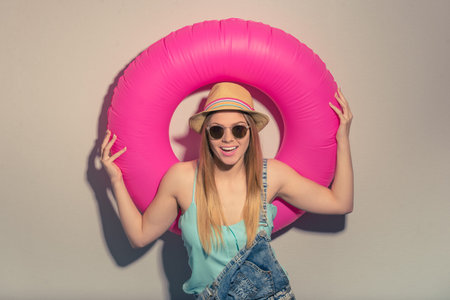Attractive girl in summer clothes and sun glasses is holding a swim ring, looking at camera and smiling, against gray backgroundの写真素材
