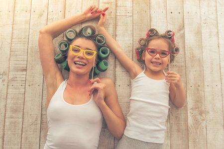 Top view of young mother and her daughter with hair curlers on their heads holding paper glasses on stick and smiling, lying on the floor at homeの写真素材