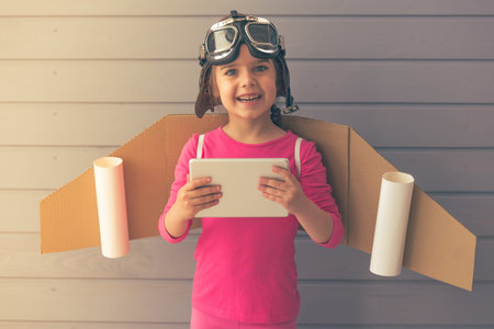 Cute little girl dressed like a pilot with toy wings is using a tablet, smiling and looking at camera, against gray wallの写真素材