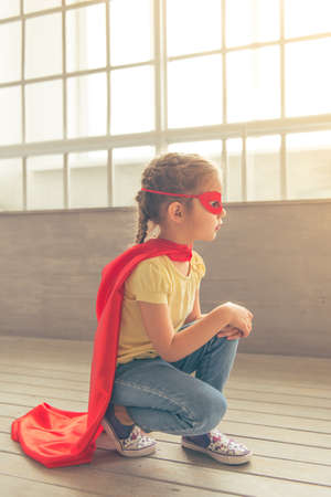 Side view of cute little girl dressed like superhero playing at home, squatting against windowsの写真素材