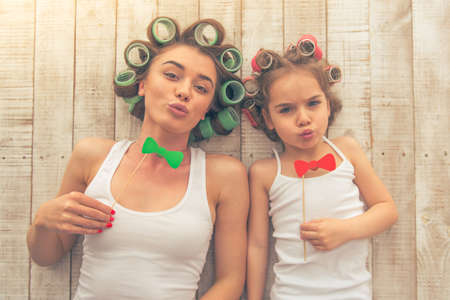 Top view of young mother and her daughter with hair curlers on their heads holding paper bows on stick and showing kisses, lying on the floor at homeの写真素材