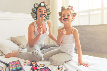 Beautiful young mother and her cute daughter in pajamas and with hair curlers are holding paper moustaches on stick and smiling while sitting on bed at homeの写真素材