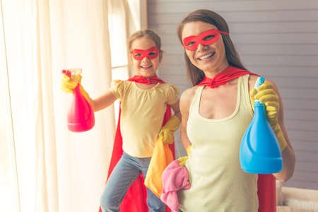 Beautiful mother and her cute little daughter dressed like superheroes are holding sprayer, looking at camera and smiling while cleaning the houseの写真素材