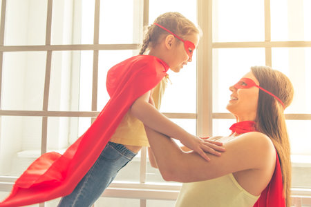 Beautiful mother and her little daughter dressed like superheroes are looking at each other and...
