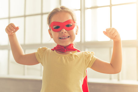 Portrait of cute little girl dressed like superhero showing her muscles, smiling and looking at camera, against windowsの写真素材