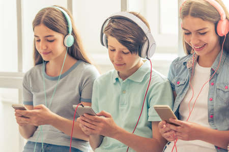Group of teenage boys and girls in headphones is listening to music using smartphones while sitting against windowの写真素材
