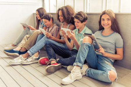 Group of teenage boys and girls is using tablets and smiling while sitting on wooden floor. One girl is looking at cameraの写真素材