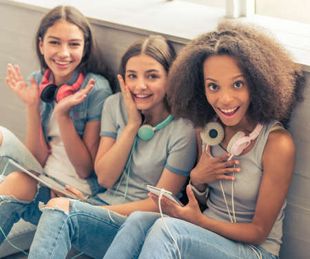Group of teenage girls with headphones and gadgets is looking at camera, showing surprise and smiling while sitting on the floorの写真素材