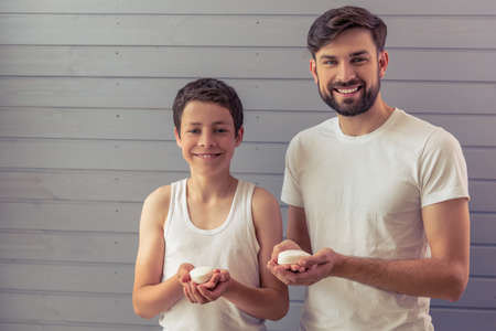 Handsome young father and his teenage son are holding soap, looking at camera and smiling, against gray wallの写真素材