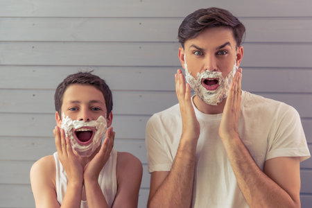 Handsome young father and his teenage son are applying  shaving foam, looking at camera and screaming, against gray wallの写真素材