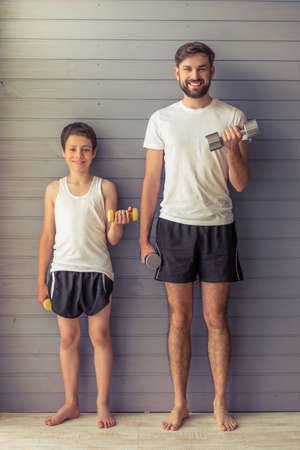 Handsome young father and his teenage son are looking at camera and smiling while working out with dumbbells at homeの写真素材