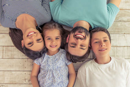 Top view of beautiful young parents, their cute little daughter and son looking at camera and smiling, lying on wooden floorの写真素材