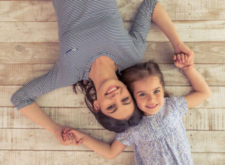 Top view of beautiful young mother and her cute little daughter holding hands, looking at camera and smiling, lying on wooden floorの写真素材