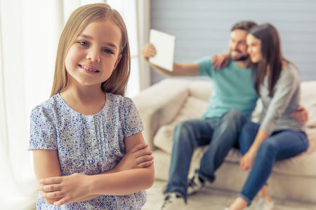 Cute little girl is looking at camera and smiling, in the background her parents are using a tablet, sitting on sofa at homeの写真素材