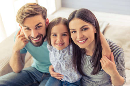 Portrait of beautiful young parents and their cute little daughter looking at camera and smiling, sitting on sofa at homeの写真素材