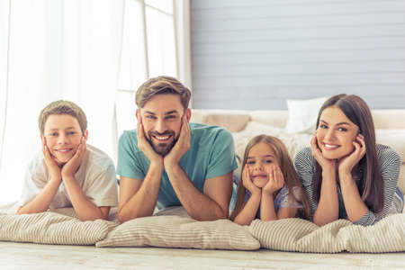 Beautiful young parents, their cute little daughter and son are looking at camera and smiling, lying on the floor at homeの写真素材