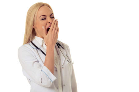 Portrait of beautiful tired female doctor in white coat covering his mouth while yawning, isolated on white backgroundの写真素材