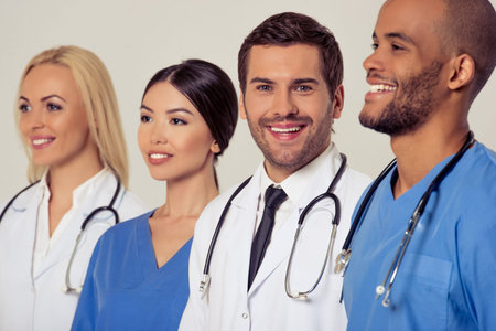 Group of medical doctors of different nationalities and genders is looking forward and smiling, standing in a row on gray background. One is looking at cameraの写真素材