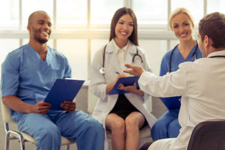 Medical doctors of different nationalities and genders are making notes, talking and smiling, sitting in office. Doctor in the foreground in focusの写真素材