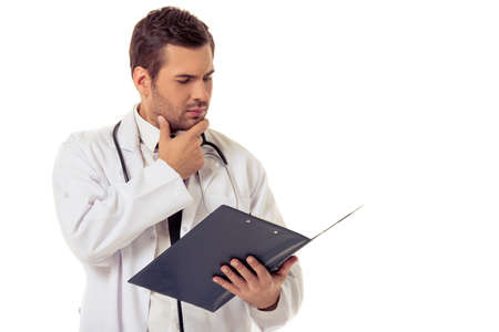 Portrait of handsome pensive doctor in white coat holding a folder and examining data, isolated on white backgroundの写真素材