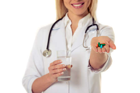 Cropped image of beautiful female doctor in white coat smiling while holding a glass of water and pills, isolated on white backgroundの写真素材