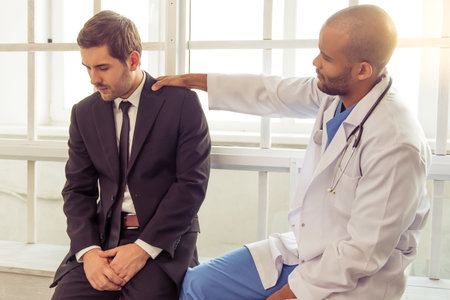Handsome Afro American doctor is keeping hand on businessman's shoulders while calming him