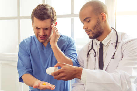 Handsome tired doctor in blue medical wear is keeping hand on his temple, having headache. Afro American doctor is giving him pillsの写真素材