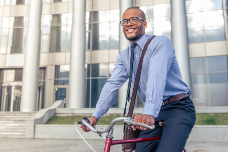 Handsome Afro American businessman in classic clothes and glasses is smiling while riding his bike outside the office buildingの写真素材