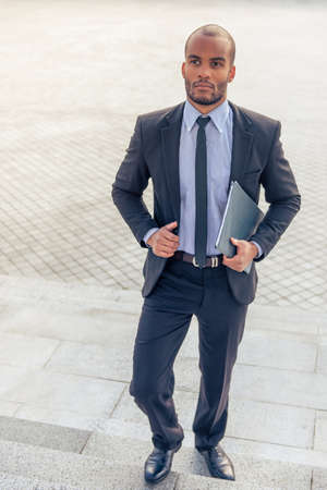 Handsome young Afro American businessman in classic suit is holding a folder and looking forward while coming upstairsの写真素材