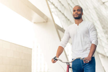 Handsome young Afro American man in casual clothes is looking forward while walking with bike outdoorsの写真素材