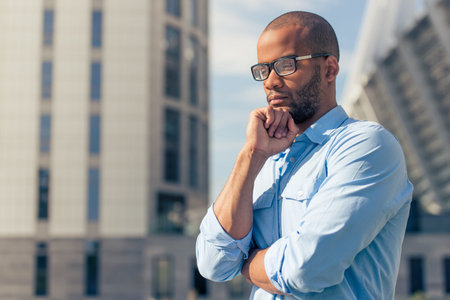 Handsome young pensive Afro American businessman in eyeglasses is thinking while standing outdoorsの写真素材