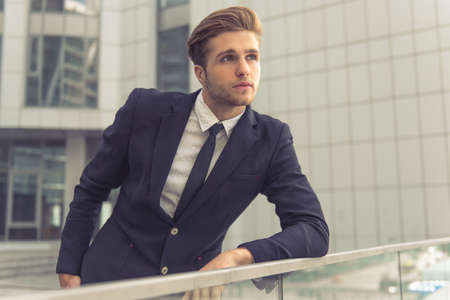 Attractive young confident businessman in classic suit is looking away, leaning on balcony balustrade, outside the office buildingの写真素材