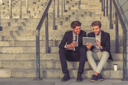 Two handsome young businessmen in formal clothes are using a tablet and smiling while sitting on stairs of the office building during coffee breakの写真素材