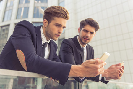 Two handsome young businessmen in classic suits are using smartphones, leaning on balcony outside the office buildingの写真素材