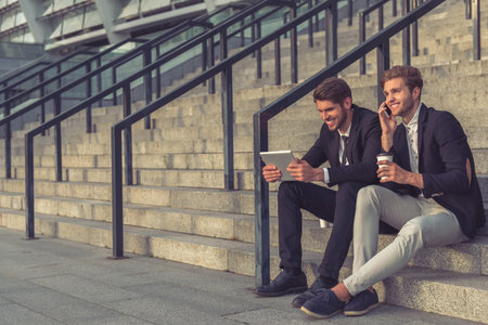 Two handsome young businessmen are using a tablet, talking on the mobile phone and smiling while...
