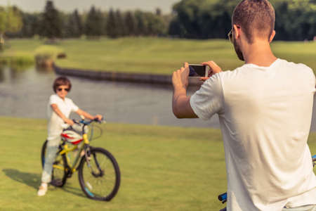 Back view of young father taking photo of his little son on bicycle, using a smart phone while they both are resting in parkの写真素材