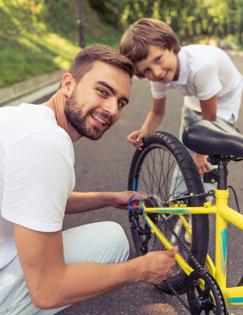 Handsome young dad and his cute little son are riding bikes in park. Both are looking at camera and smiling while father is examining his son's bicycleの写真素材