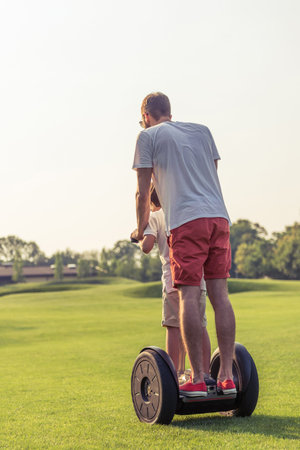 Back view of little boy and his father in casual clothes riding a segway in the park