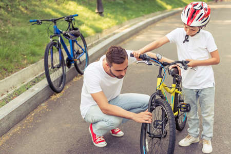 Handsome young dad and his cute little son are riding bikes in park. Father is examining his son's bicycleの写真素材