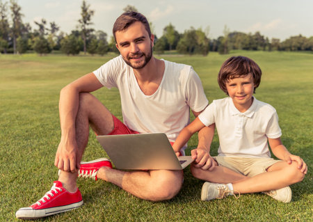 Cute little boy and his handsome young dad are using a laptop, looking at camera and smiling while sitting on the grass in the parkの写真素材