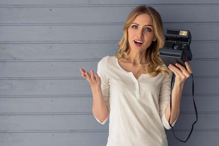 Beautiful young woman is keeping a camera and showing surprise, standing against gray wallの写真素材