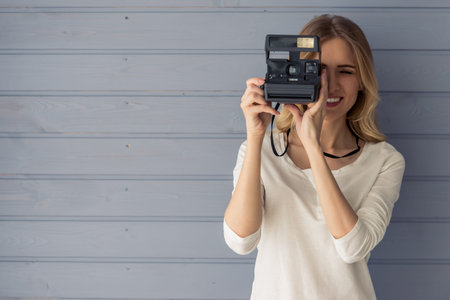 Beautiful young woman is making photo using camera and smiling, standing against gray wall