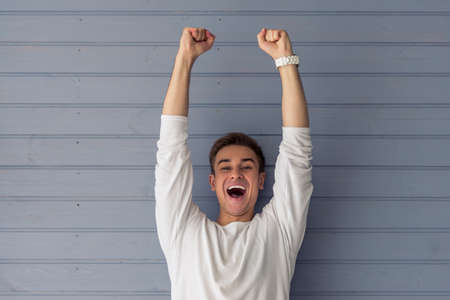 Handsome young man in casual clothes is keeping hands in fists up, looking at camera and screaming with happiness, standing against gray wallの写真素材