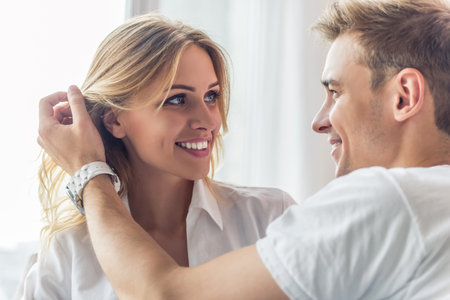 Portrait of beautiful young couple looking at each other and smiling while sitting at home. Man is...